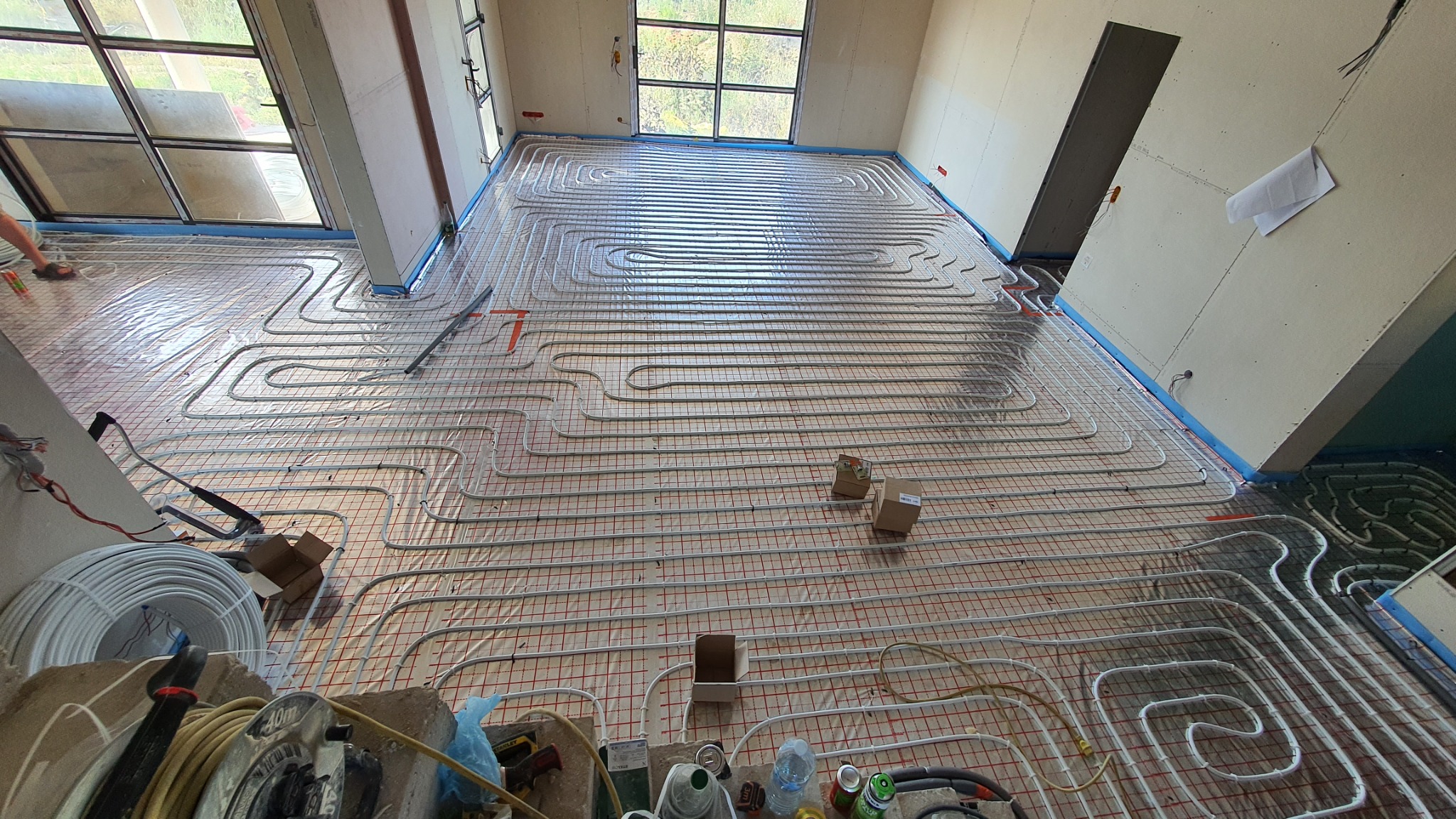 Rozłożona instalacja ogrzewania podłogowego w nowym budynku, rury ułożone w pętle na siatce, widoczne narzędzia i materiały budowlane, surowe ściany i okna.