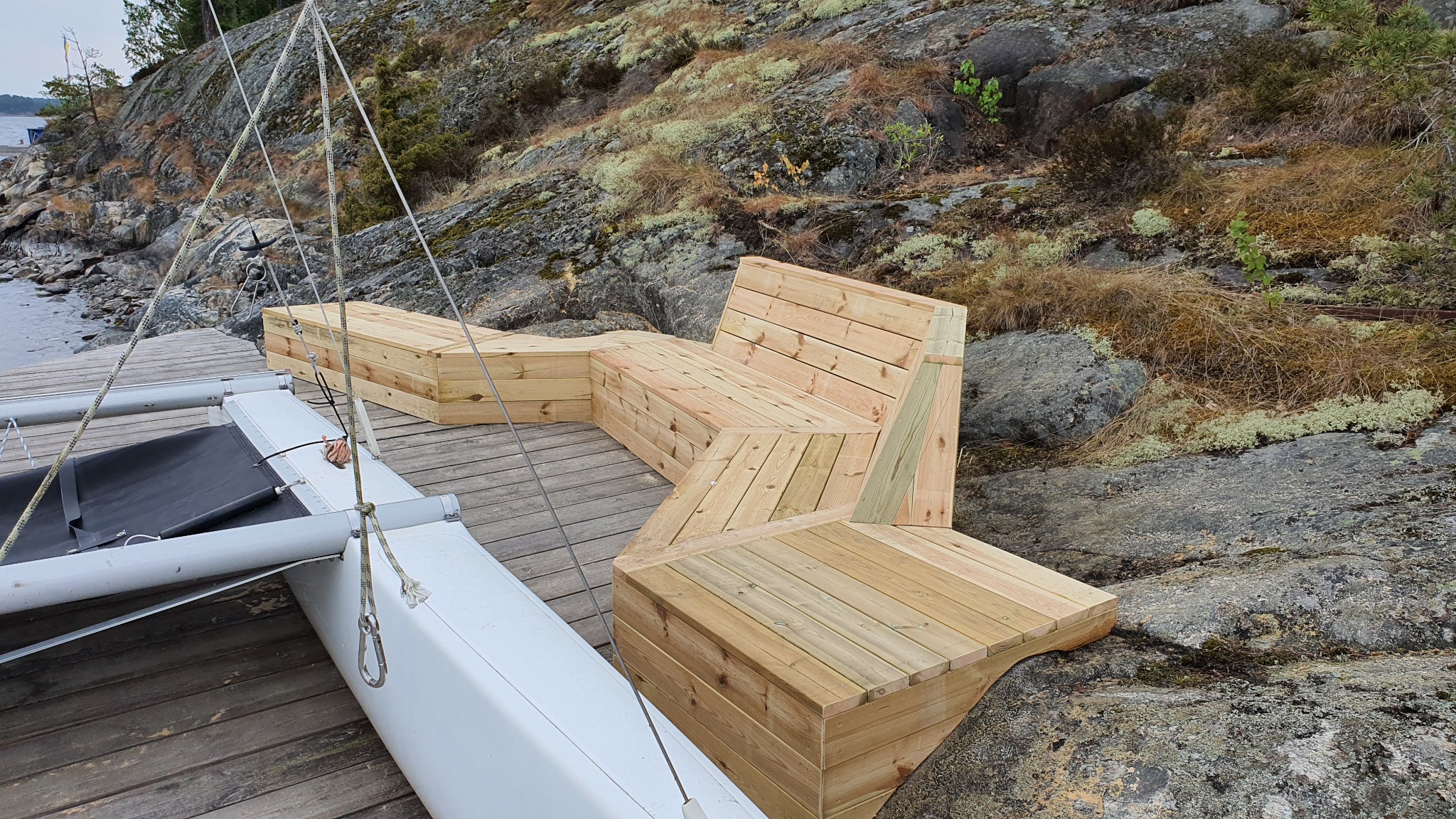 Nowoczesna, wieloelementowa ławka z jasnego drewna na drewnianym pomoście, obok katamaranu, z skalistym brzegiem w tle porośniętym mchem i roślinnością.