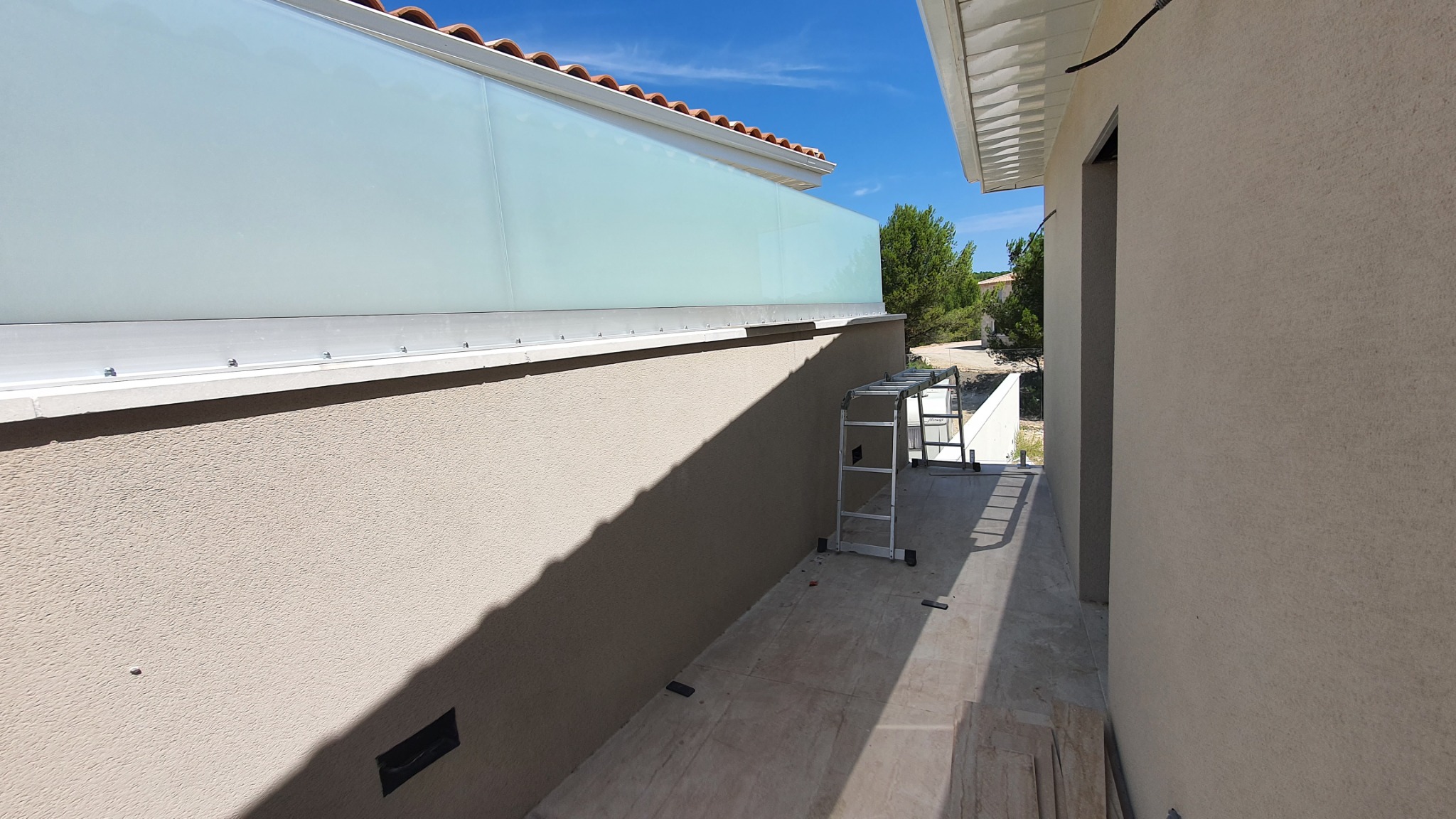 Zewnętrzna ściana budynku z zamontowaną szklaną balustradą, widoczny fragment elewacji i drabina aluminiowa.