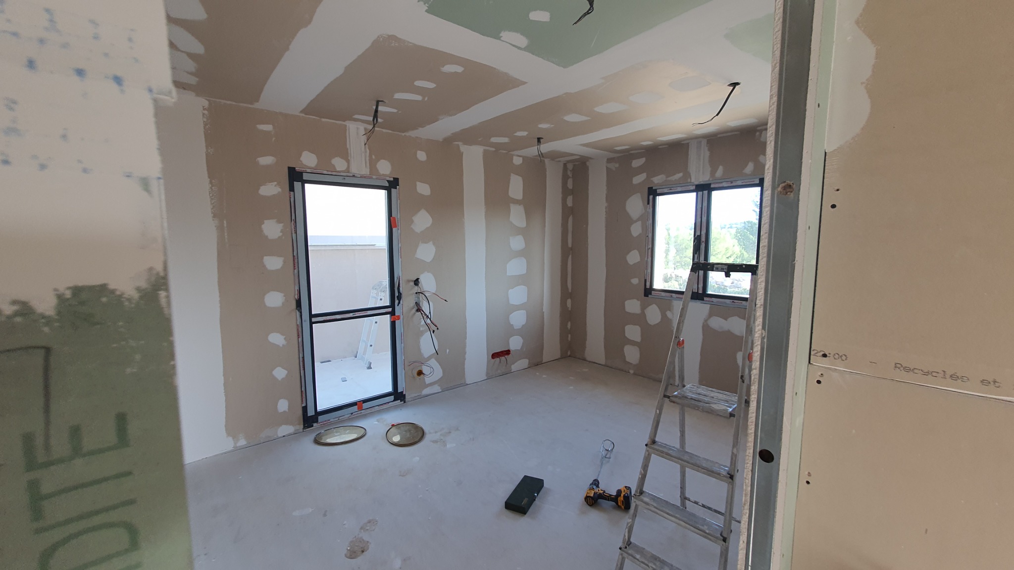Pokój w trakcie montażu płyt kartonowo-gipsowych na ścianach i suficie, widoczne narzędzia, drabina, okno i drzwi balkonowe.