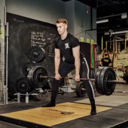 Mężczyzna w czarnym stroju sportowym przygotowuje się do podniesienia sztangi w siłowni, widoczne obciążenia, tablica z zapisanym planem treningowym oraz osoba w tle.
