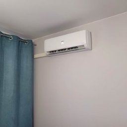 OPTIBUD SP. ZO.O. - Biały klimatyzator ścienny zamontowany w pokoju z jasnymi ścianami i niebieską zasłoną po lewej stronie.