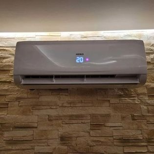 Biały klimatyzator marki Heiko z wyświetlaczem temperatury 20 stopni Celsjusza, zamontowany na ścianie wyłożonej kamienną okładziną w jednym z domów w Lublinie. Widoczne oświetlenie sufitowe.