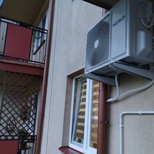 Zewnętrzna jednostka klimatyzacji marki Heiko zamontowana na ścianie budynku, widoczne rury instalacyjne i okno z zasłoną w tle.