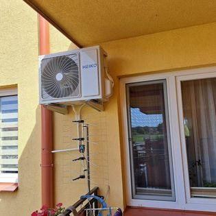 Biała jednostka zewnętrzna klimatyzacji marki HEIKO zamontowana na żółtej elewacji budynku z widoczną anteną telewizyjną i oknem z odbiciem krajobrazu.