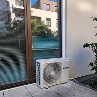 Biała jednostka zewnętrzna klimatyzatora marki Heiko, umieszczona na szarym tarasie przed dużym oknem z żaluzjami, z widocznym odbiciem budynku w szybie.