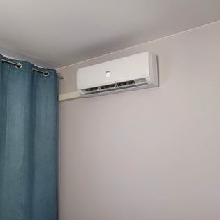 Biały klimatyzator ścienny zamontowany w pokoju z jasnymi ścianami i niebieską zasłoną po lewej stronie.