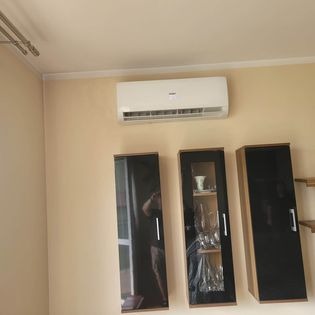 Biały klimatyzator ścienny zamontowany w pomieszczeniu z beżowymi ścianami, widoczne trzy czarne szafki wiszące z chromowanymi uchwytami.