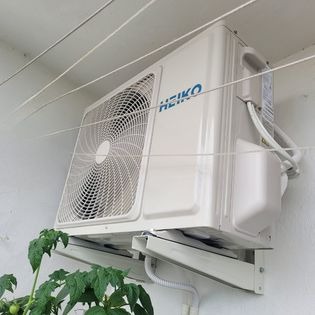 Biała jednostka zewnętrzna klimatyzacji marki HEIKO zamontowana na wspornikach na białej ścianie, z widoczną rośliną w lewym dolnym rogu i sznurkami do suszenia prania u góry.