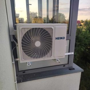 Biała jednostka zewnętrzna klimatyzatora marki Heiko zamontowana na wspornikach w oknie balkonowym, widok z zewnątrz, z zielenią w tle.