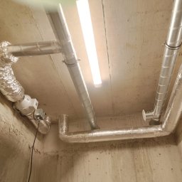 ZALST STEFAN STADNIK - Montaż rekuperacji: kanały wentylacyjne z folii aluminiowej przymocowane do sufitu w surowym wnętrzu, widoczny wentylator i lampa sufitowa.