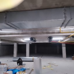 ZALST STEFAN STADNIK - Montaż kanałów wentylacyjnych i rur pod sufitem w surowym wnętrzu. Widoczne elementy instalacji i materiały budowlane. Dwa wentylatory kanałowe.