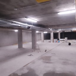 ZALST STEFAN STADNIK - Surowe wnętrze hali z widocznymi elementami instalacji wentylacyjnej pod sufitem, drabina i przewody na posadzce, betonowe ściany i filary.