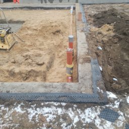 ZALST STEFAN STADNIK - Fundamenty z pionowymi rurami kanalizacyjnymi w wykopie, częściowo zasypanym piaskiem. Widoczny pracownik w pomarańczowym kombinezonie i ubijarka wibro.