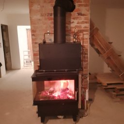 ZALST STEFAN STADNIK - Czarny piec wolnostojący z płomieniami w szklanej obudowie, z instalacją miedzianą na tle surowej cegły w remontowanym wnętrzu.