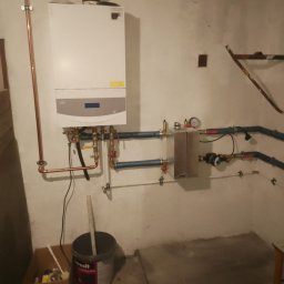 ZALST STEFAN STADNIK - Biały piec gazowy zamontowany na ścianie z widocznymi rurami miedzianymi i instalacją hydrauliczną. Obok stoi wiadro z narzędziami.