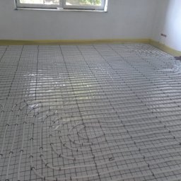 ZALST STEFAN STADNIK - Białe rury ogrzewania podłogowego ułożone na siatce w nowym pomieszczeniu, zabezpieczone taśmą malarską przy ścianie i z widocznymi rurami doprowadzającymi.