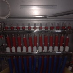 ZALST STEFAN STADNIK - Rozdzielacz ogrzewania podłogowego w metalowej szafce, z czerwonymi i niebieskimi rurami. Widoczne zawory i odpowietrzniki. Instalacja w nowym budynku.
