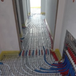 ZALST STEFAN STADNIK - Instalacja ogrzewania podłogowego z rurami w kolorach niebieskim i czerwonym, rozłożonymi na metalowej siatce w korytarzu podczas prac wykończeniowych.
