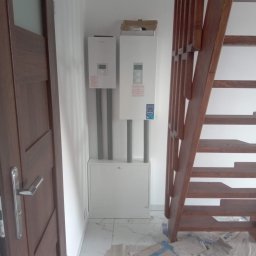 ZALST STEFAN STADNIK - Dwa białe piece gazowe zamontowane na ścianie w Porębie, z widocznymi rurami i szafką pod nimi. Obok drewniane schody i drzwi. Podłoga wyłożona marmurowymi płytkami.