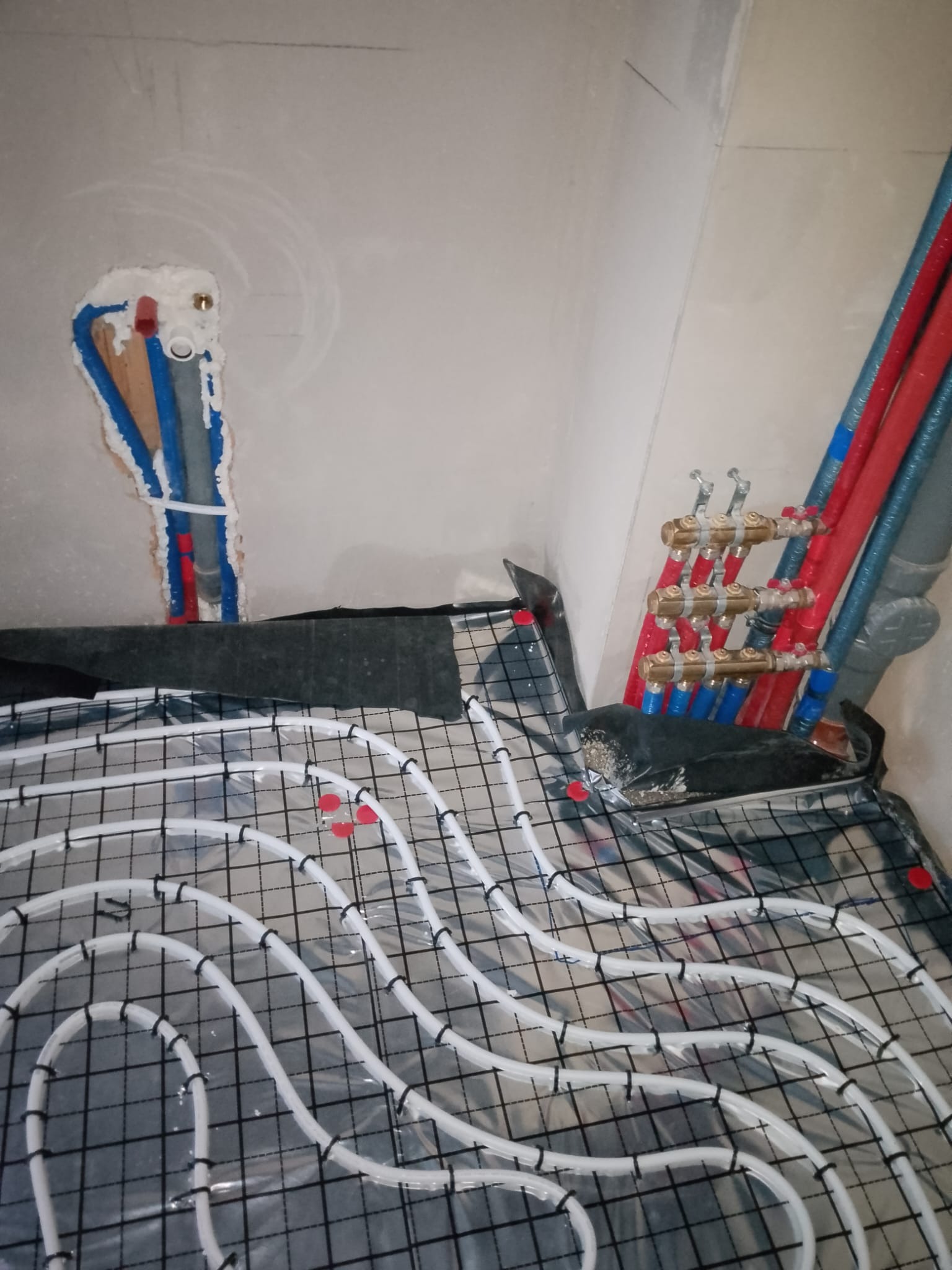 Białe rury ogrzewania podłogowego ułożone na czarnej siatce, z czerwonymi i niebieskimi rurami w ścianie i rozdzielaczem. Etap instalacji w surowym wnętrzu.