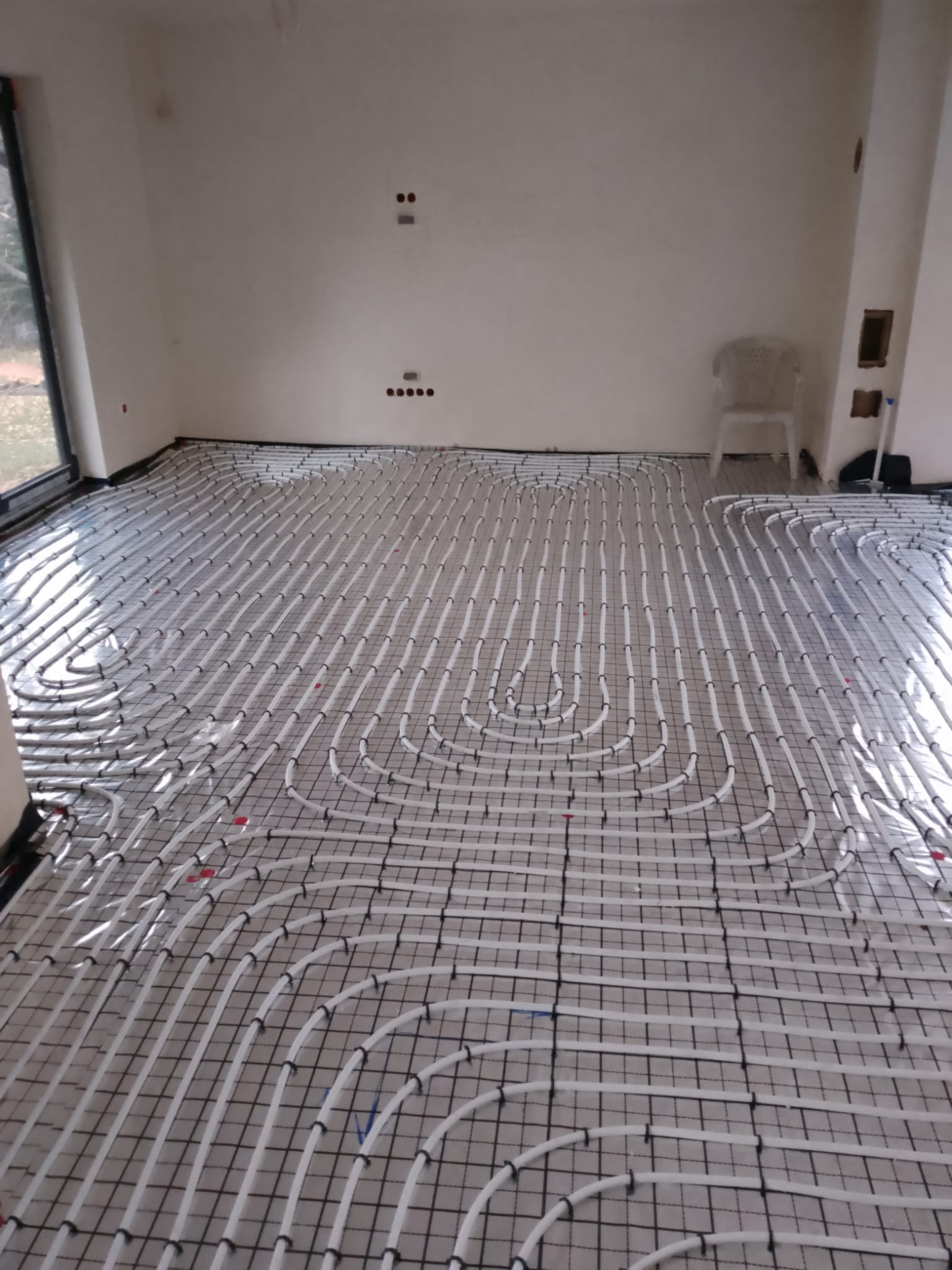 Białe rury ogrzewania podłogowego ułożone na siatce w surowym wnętrzu z oknem. Widoczne punkty instalacji elektrycznej na ścianie.