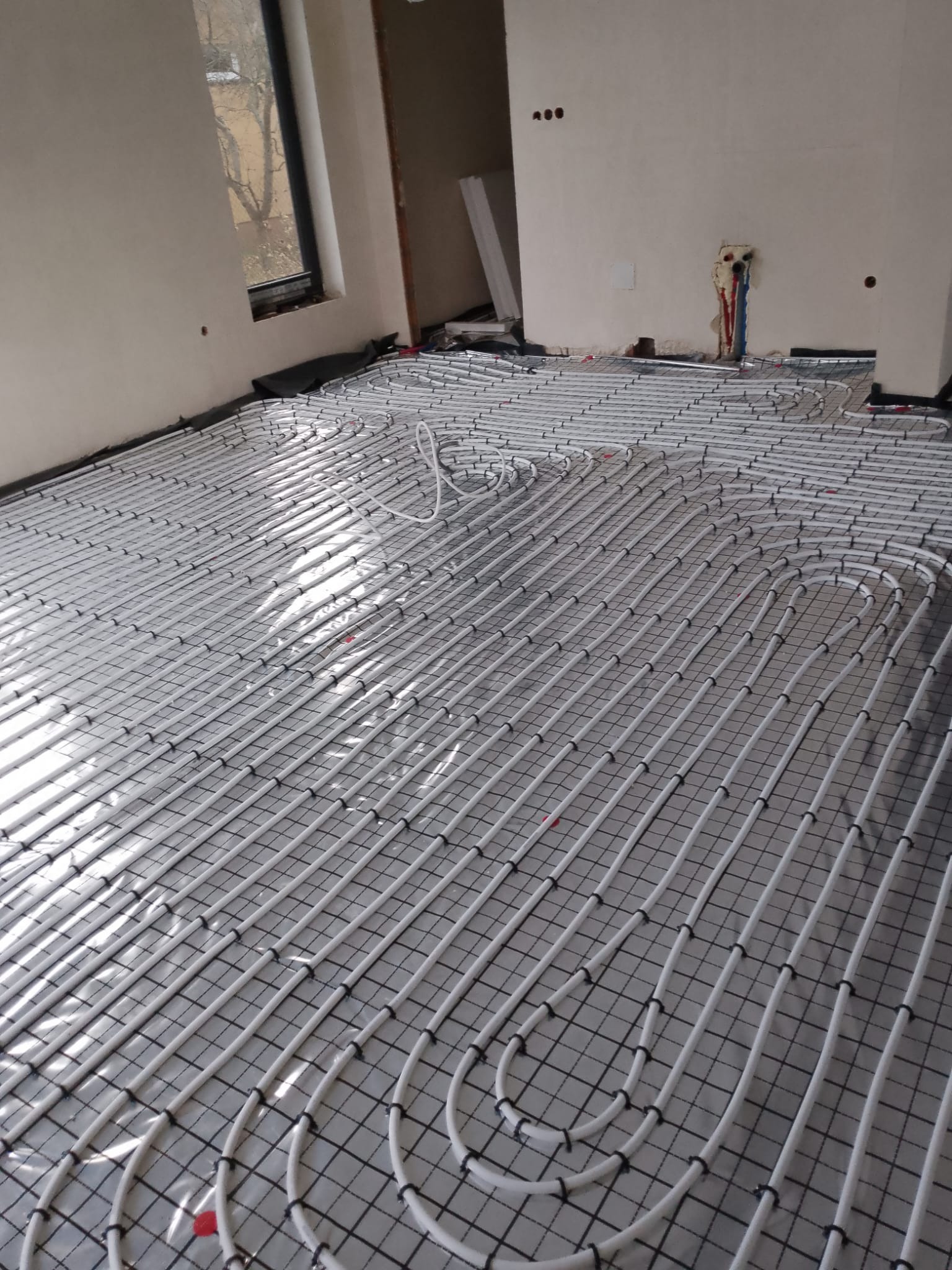 Białe rury ogrzewania podłogowego ułożone na czarnej siatce w surowym pomieszczeniu z oknem. Widoczne wystające rury instalacji.