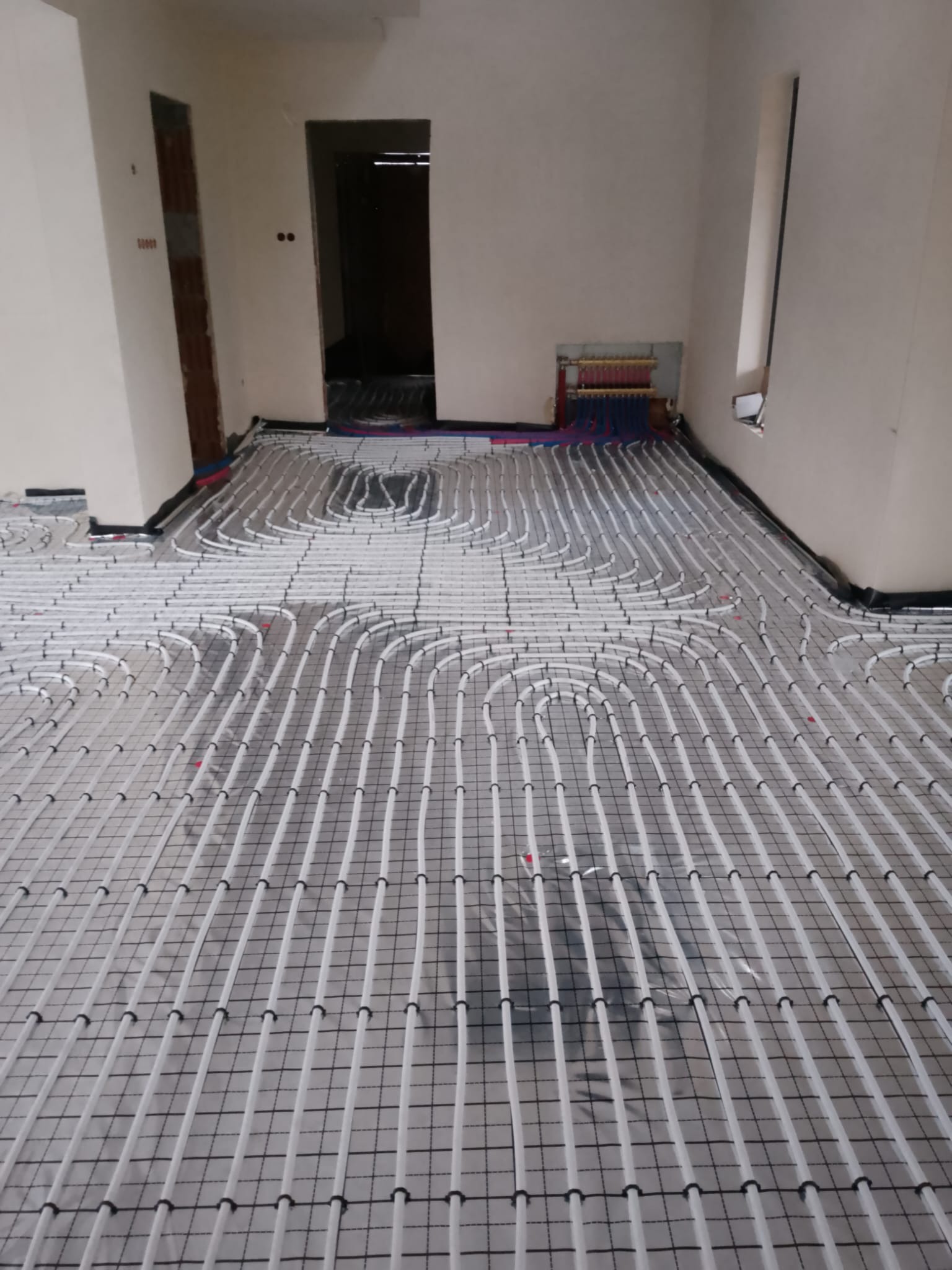 Białe rury ogrzewania podłogowego ułożone na czarnej folii w jasnym pomieszczeniu. Widoczny rozdzielacz z rurami w kolorach czerwonym i niebieskim.