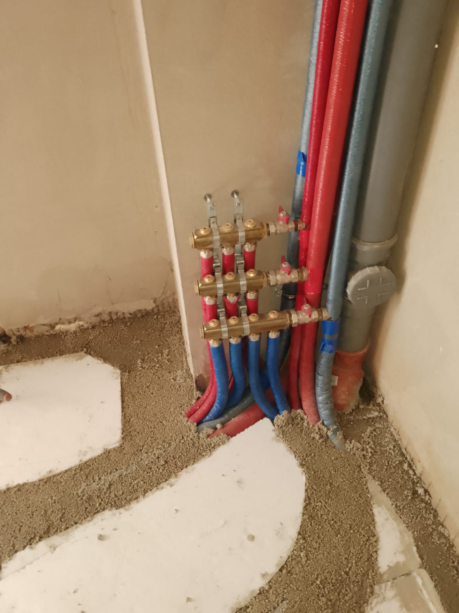 Rozdzielacz ogrzewania podłogowego z rurami PEX w kolorze czerwonym i niebieskim, zamontowany na ścianie, częściowo zalany wylewką. Widoczne elementy instalacji.