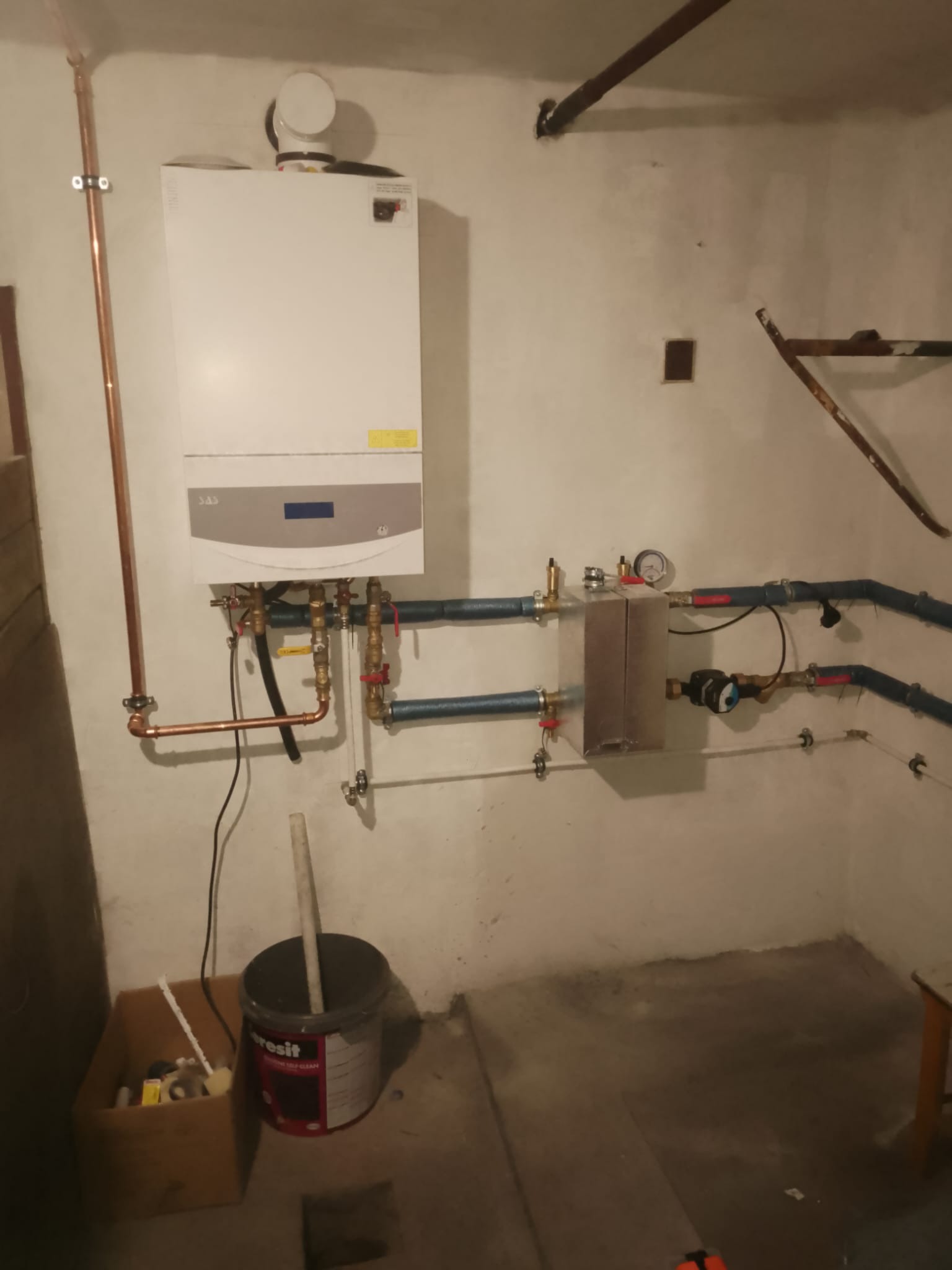 Biały piec gazowy zamontowany na ścianie z widocznymi rurami miedzianymi i instalacją hydrauliczną. Obok stoi wiadro z narzędziami.