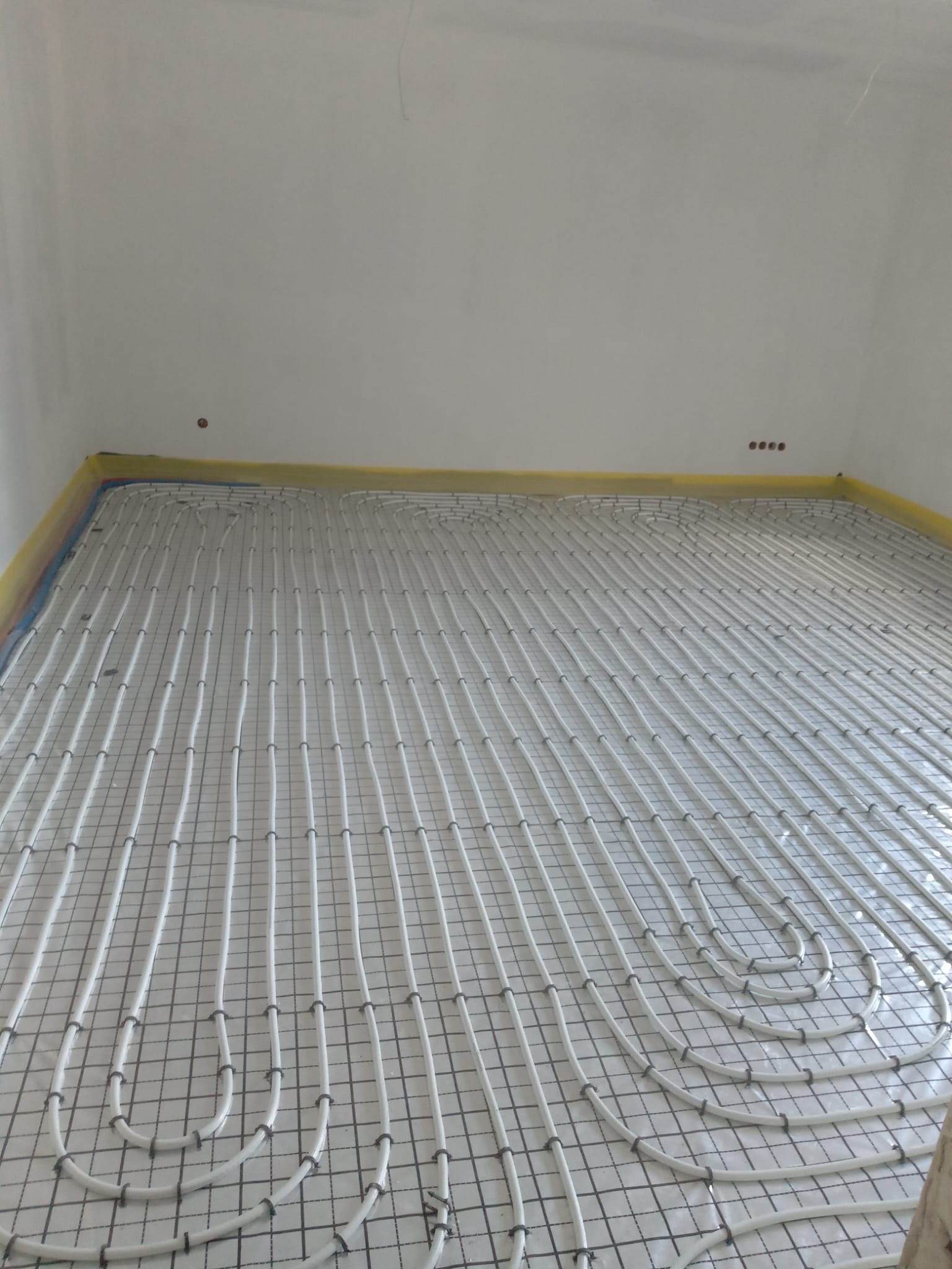 Białe rury ogrzewania podłogowego ułożone na siatce w pomieszczeniu z żółtą taśmą przy ścianach. Widok z góry ukazuje regularny wzór instalacji.