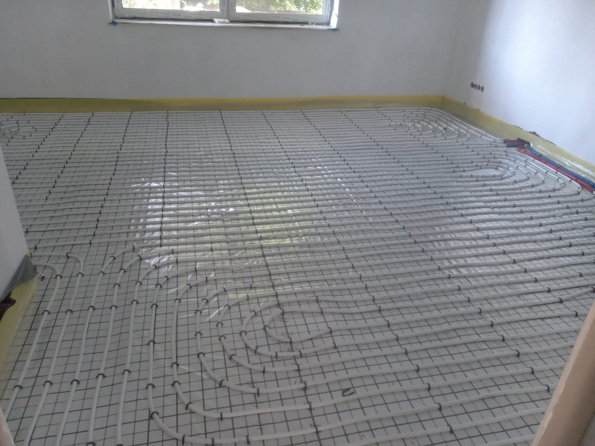 Białe rury ogrzewania podłogowego ułożone na siatce w nowym pomieszczeniu, zabezpieczone taśmą malarską przy ścianie i z widocznymi rurami doprowadzającymi.
