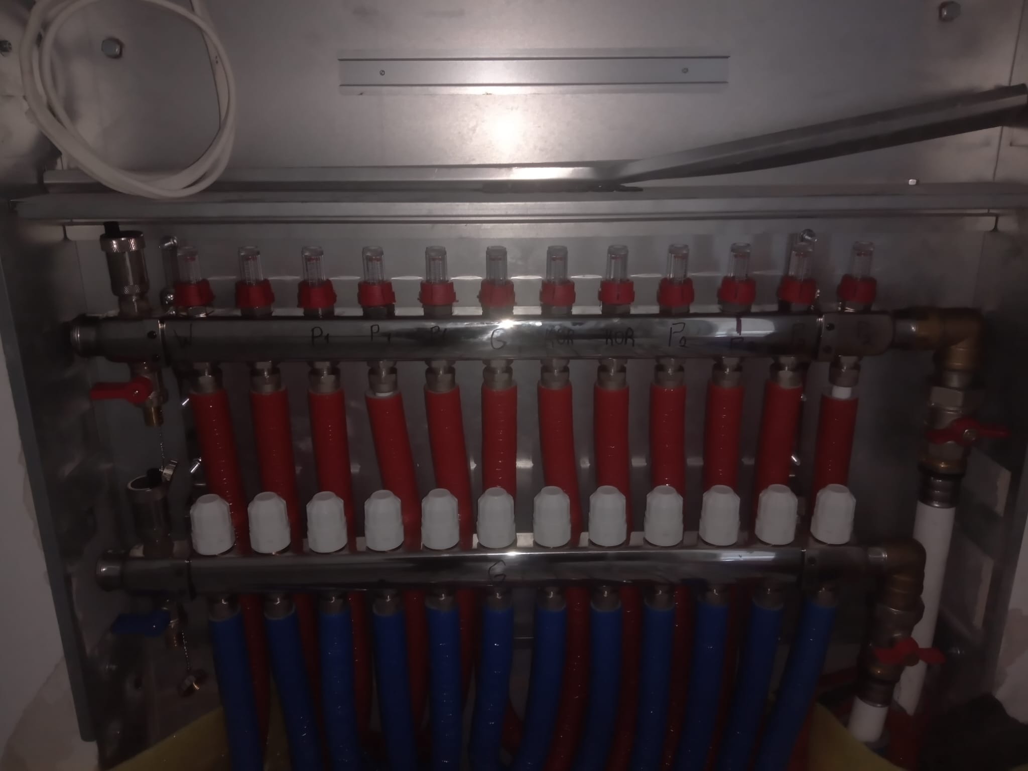Rozdzielacz ogrzewania podłogowego w metalowej szafce, z czerwonymi i niebieskimi rurami. Widoczne zawory i odpowietrzniki. Instalacja w nowym budynku.