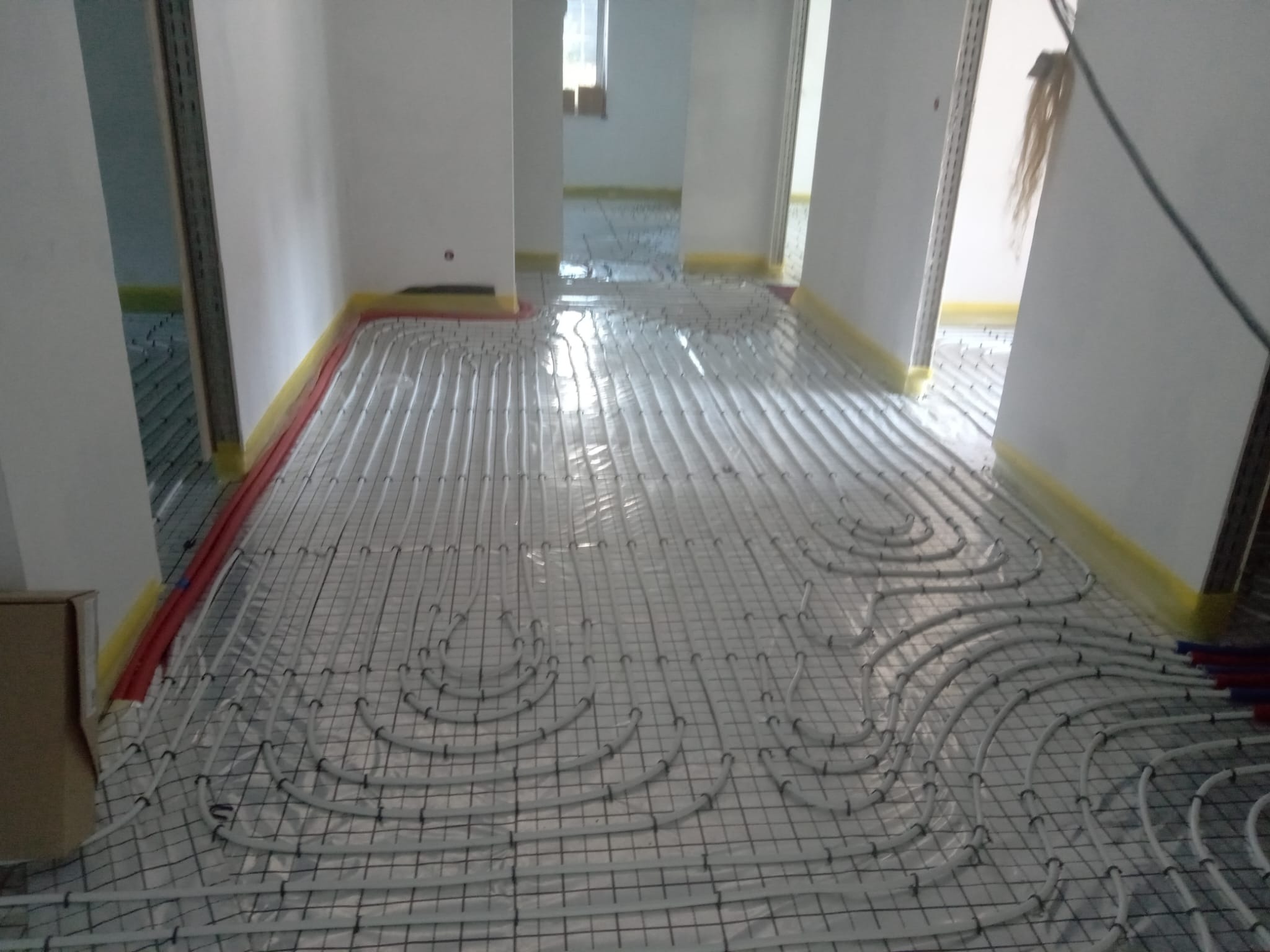 Białe rury ogrzewania podłogowego ułożone na siatce w jasnym pomieszczeniu z przygotowanymi ścianami, widok z góry na rozłożoną instalację przed wylaniem posadzki.