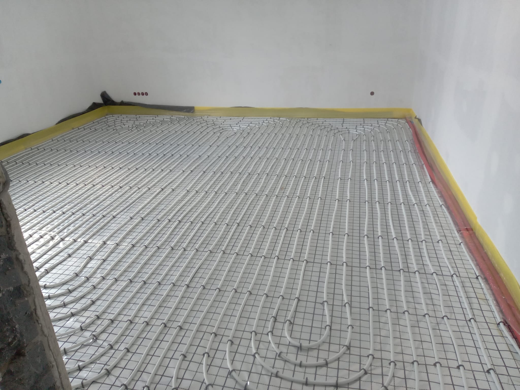 Białe rury ogrzewania podłogowego ułożone na siatce w jasnym pomieszczeniu, przygotowane do zalania posadzką. Widoczne zabezpieczenia ścian folią.