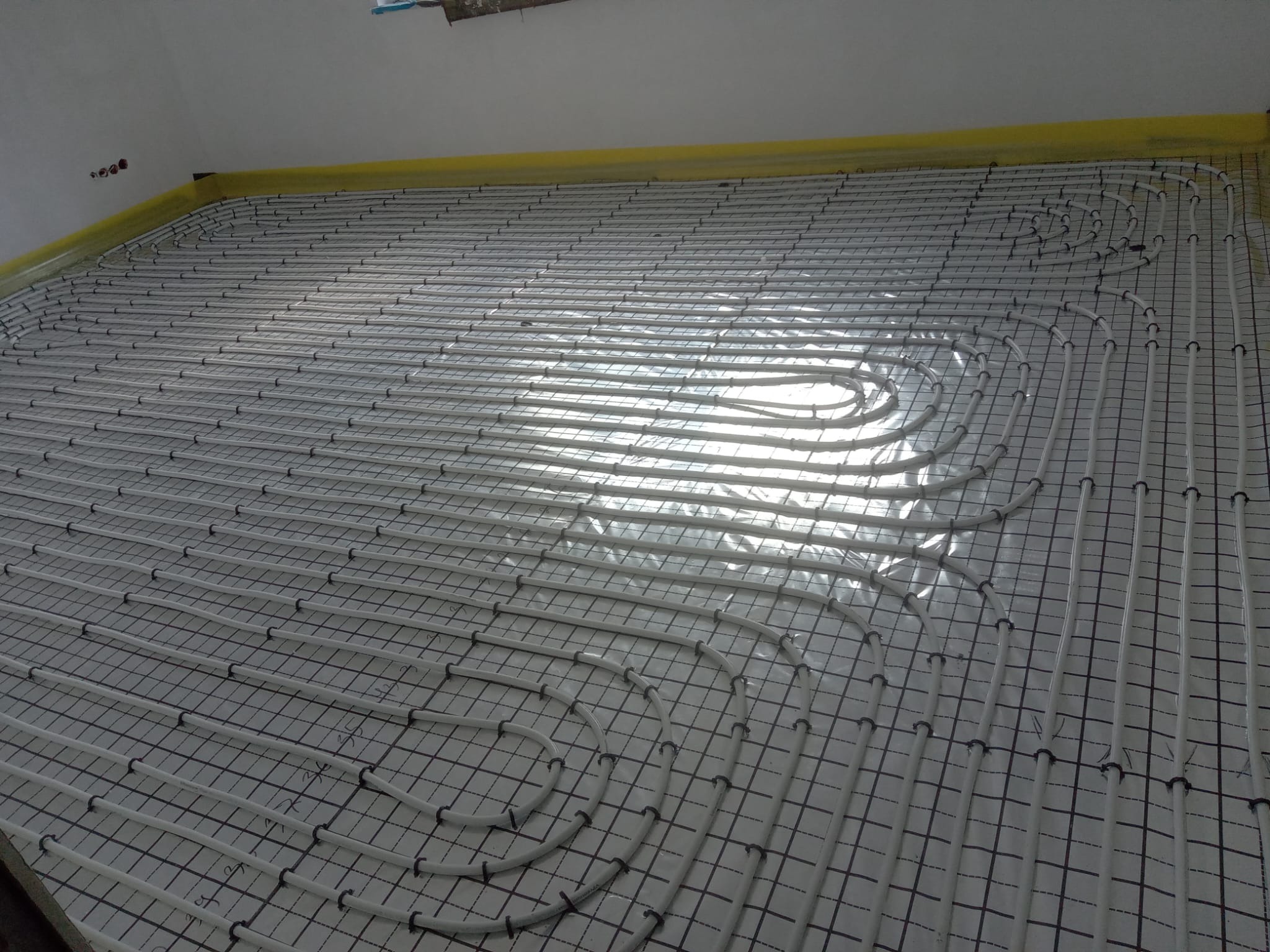 Białe rury ogrzewania podłogowego ułożone spiralnie na czarnej siatce, zabezpieczone żółtą folią przy ścianie. Widoczne złącza rur. Jasne światło odbija się od powierzchni.