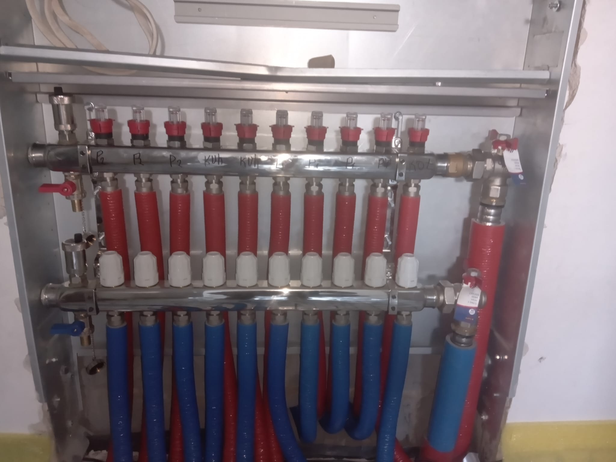 Rozdzielacz ogrzewania podłogowego z rurami w kolorze niebieskim i czerwonym, zamontowany w metalowej szafce, widok z bliska.