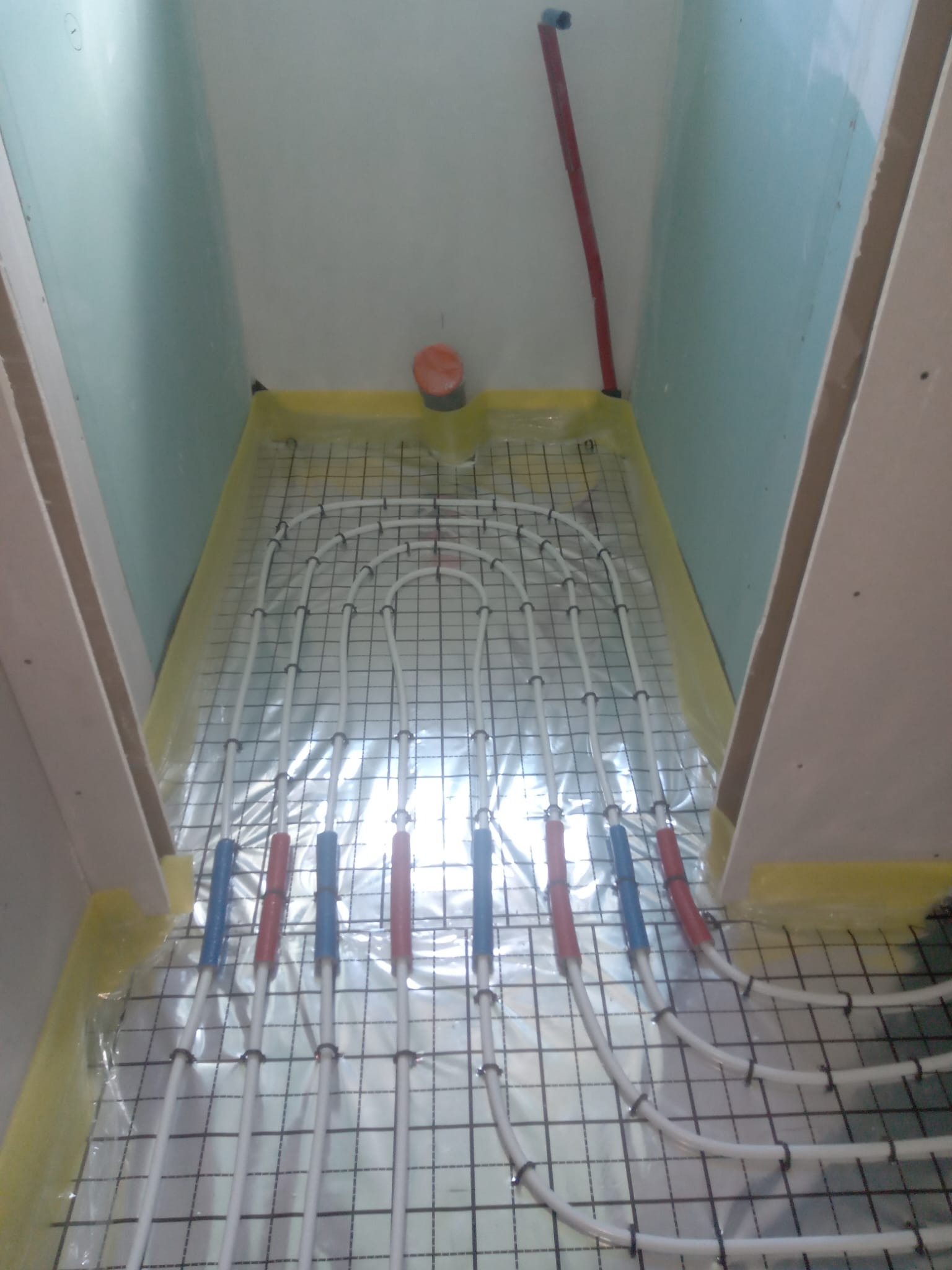 Białe rury ogrzewania podłogowego w łazience, zamocowane do czarnej siatki, widok z góry. Rury z czerwonymi i niebieskimi oznaczeniami.