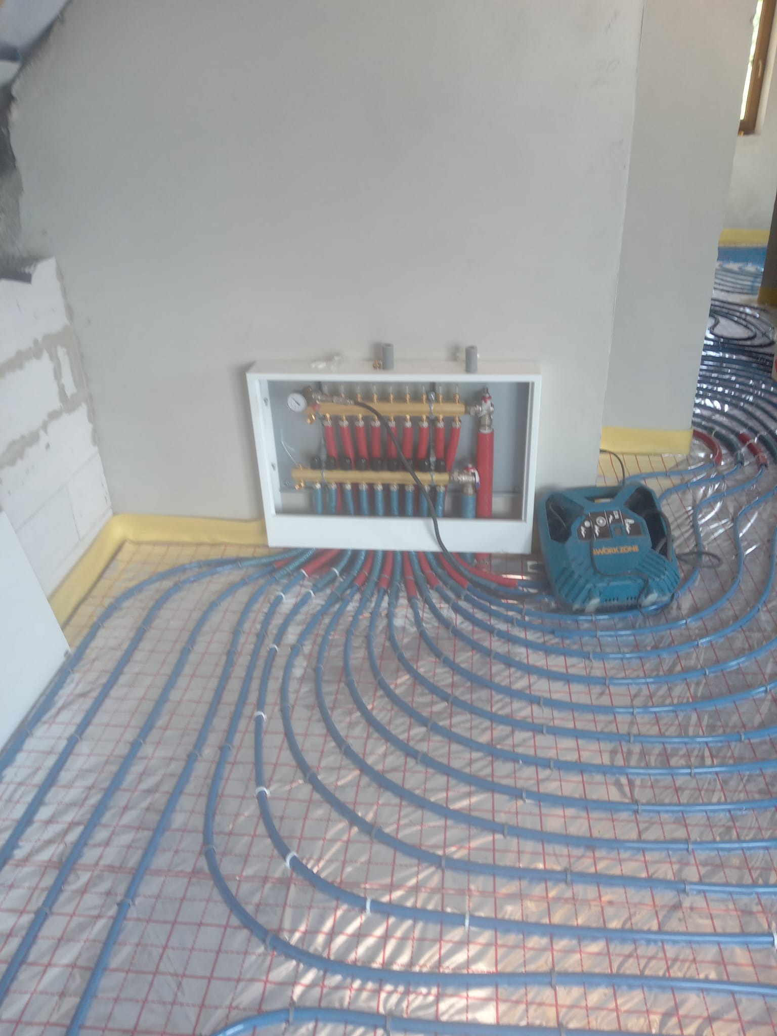 Rozłożone rury ogrzewania podłogowego na izolacji, z podłączonym rozdzielaczem w białej skrzynce i narzędziem do montażu obok. Widok z góry, surowy stan.
