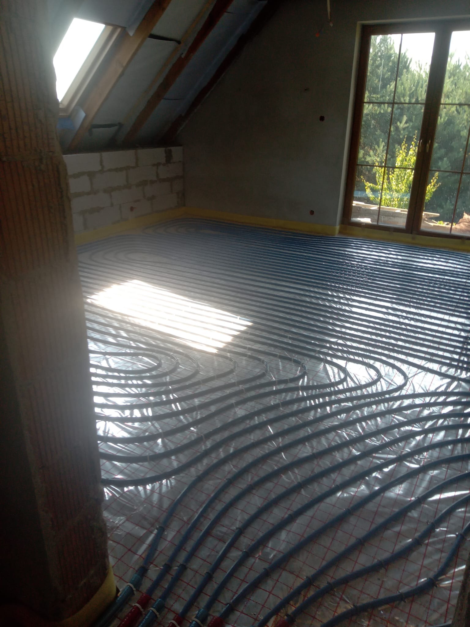 Niebieskie rury ogrzewania podłogowego ułożone na folii, widok z góry na poddaszu z oknem dachowym i dużym oknem z widokiem na zieleń.