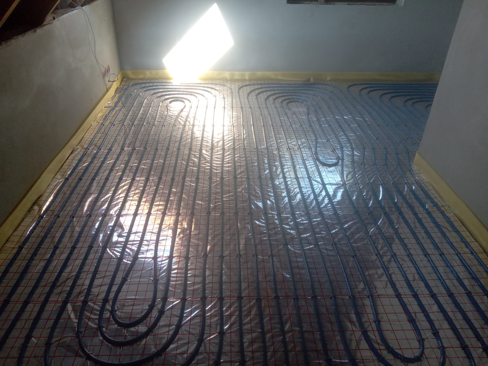 Instalacja ogrzewania podłogowego z niebieskich rur ułożonych na folii izolacyjnej, widok z góry, jasne światło dzienne wpadające przez okno.