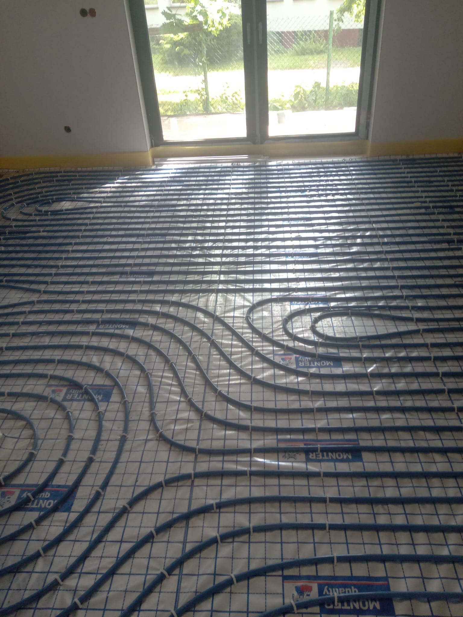 Niebieskie rury ogrzewania podłogowego ułożone na folii z siatką, widoczne okno i przygotowane otwory w ścianie.