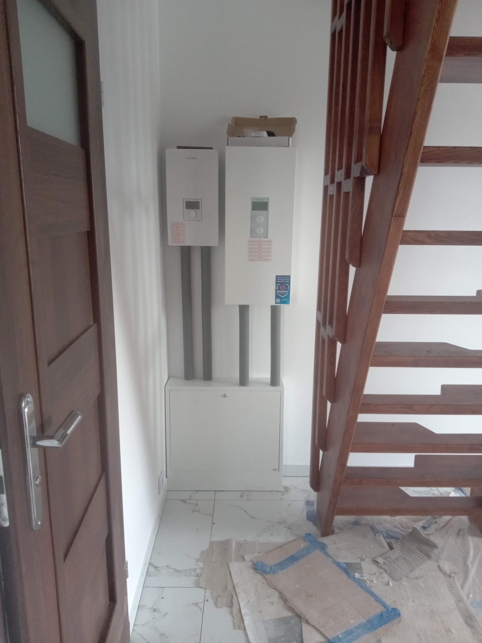 Dwa białe piece gazowe zamontowane na ścianie w Porębie, z widocznymi rurami i szafką pod nimi. Obok drewniane schody i drzwi. Podłoga wyłożona marmurowymi płytkami.