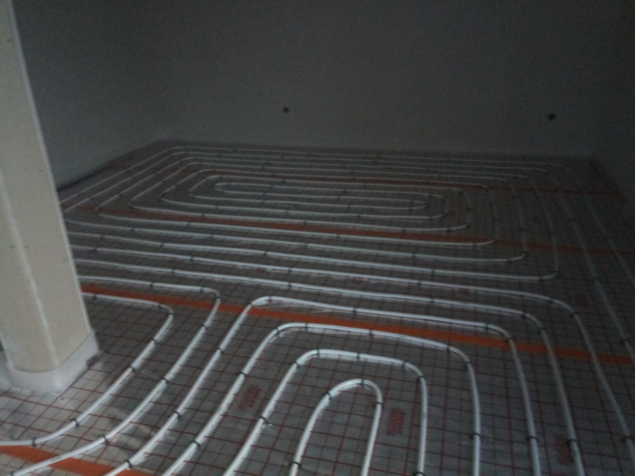 Rozłożona instalacja ogrzewania podłogowego z rurkami przymocowanymi do siatki, widok z góry na podłogę w trakcie montażu w pomieszczeniu.