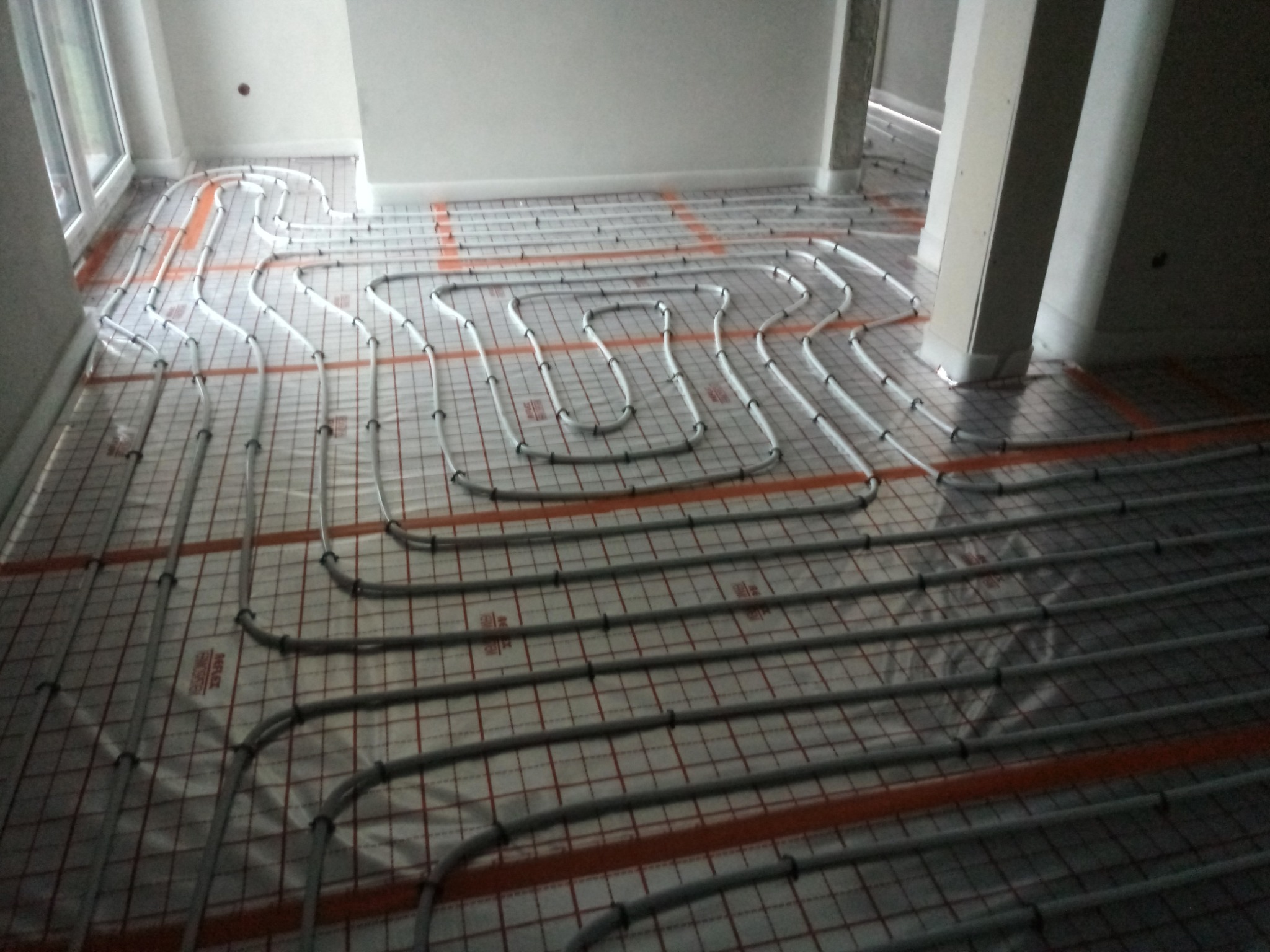 Rozłożone rury ogrzewania podłogowego na folii z siatką w jasnym pomieszczeniu, widoczne pionowe słupy.