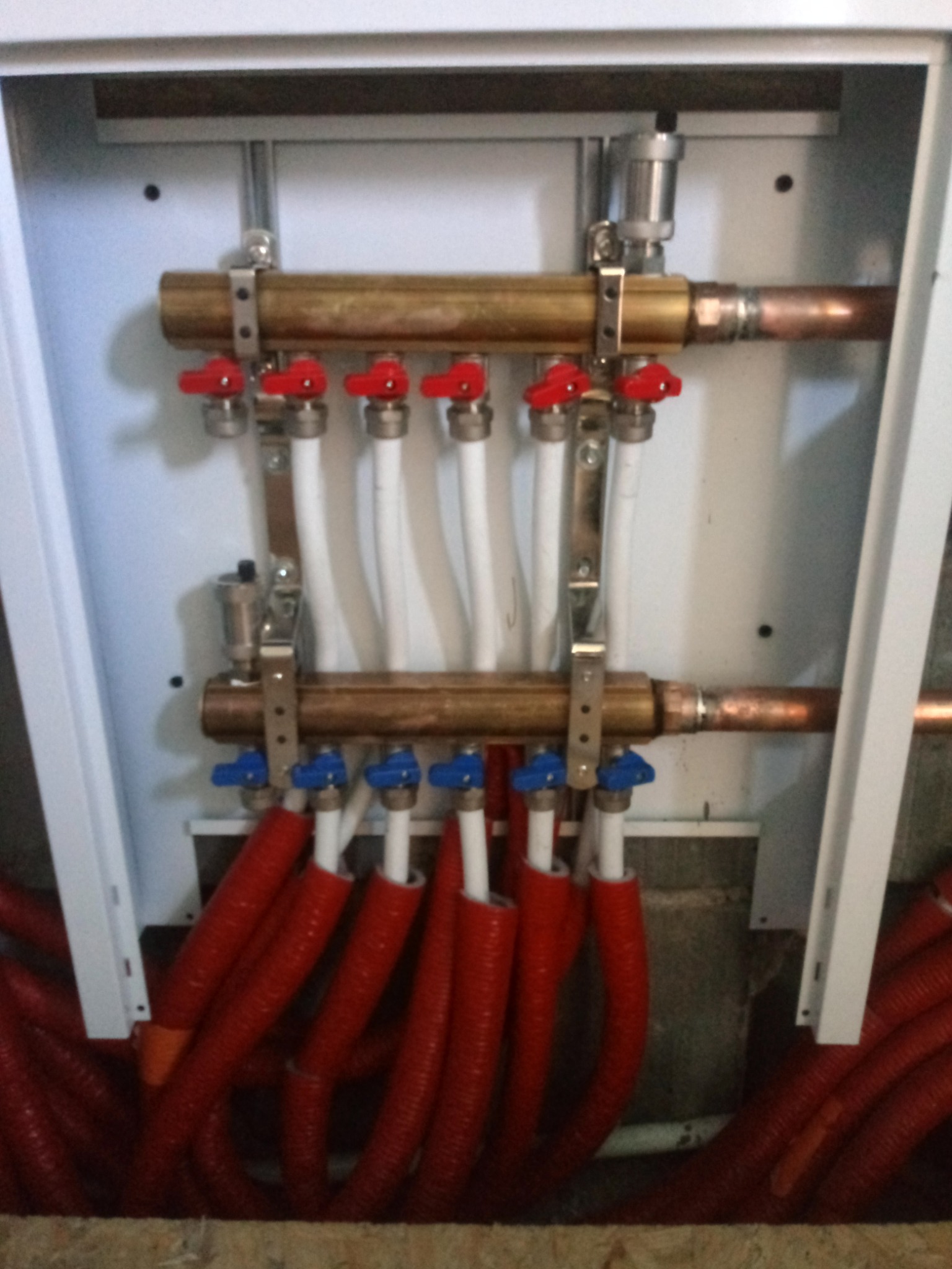 Rozdzielacz ogrzewania podłogowego w szafce, z rurami w izolacji termicznej w kolorze czerwonym i zaworami z czerwonymi i niebieskimi pokrętłami.