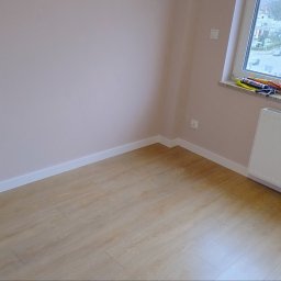 DANIEL - Wnętrze pokoju po remoncie: świeżo pomalowane ściany na delikatny róż, jasne panele podłogowe, białe listwy przypodłogowe i grzejnik pod oknem z narzędziami na parapecie.