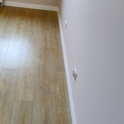 DANIEL - Jasne panele podłogowe ułożone wzdłuż ściany pomalowanej na delikatny róż, z widoczną białą listwą przypodłogową i gniazdkami elektrycznymi.