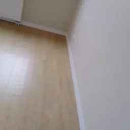 Świeżo wyremontowane wnętrze z panelami podłogowymi w odcieniach jasnego drewna, pomalowane na biało ściany z listwami przypodłogowymi, okno z grzejnikiem oraz gniazdko elektryczne.