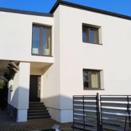 Dwupiętrowy dom z białą elewacją i ciemnymi oknami, widoczna antena satelitarna i fragment samochodu dostawczego z narzędziami.